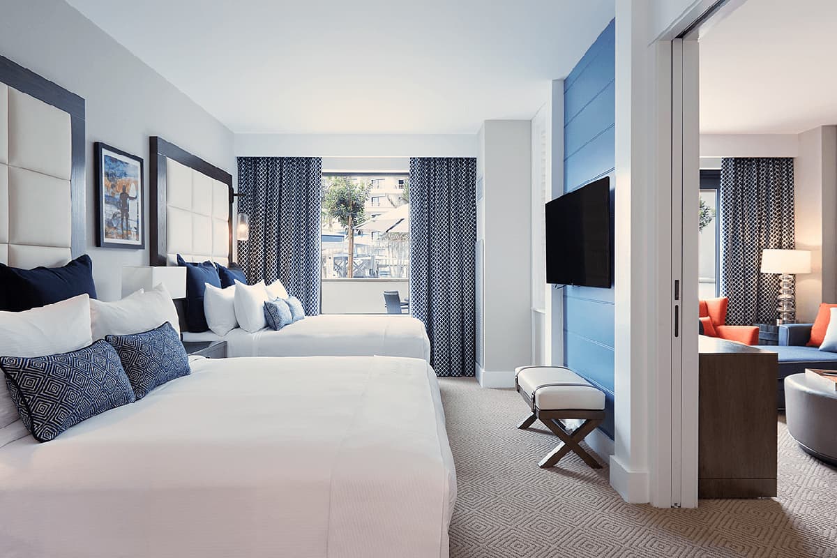 A bright, modern hotel suite with a coastal theme featuring two queen beds made up with crisp white linens and accented with navy blue and patterned throw pillows.